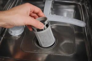 A hand removing cylindrical mesh filter from inside a stainless steel dishwasher.